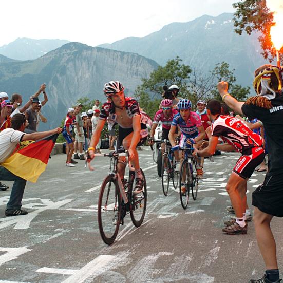 Cykelrytteren Fränk Schleck cykler på Alpe d'Huez under Tour de France 2006. Til højre ses jublende tilskuere. Én af dem med et tændt romerlys i hånden. Bag Schleck ses yderligere cykelryttere - Damiano Cunego og Eddy Mazzoleni.