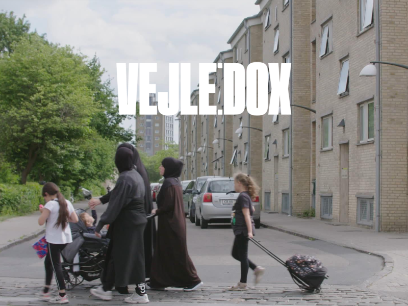 Stillbillede fra filmen som viser mennesker med kufferter foran boligblok.