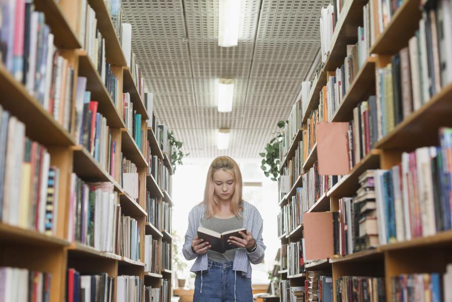 En pige kigger eller læser i en bog. Hun står mellem to bogreoler fyldt med bøger. 